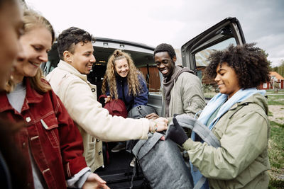 Smiling woman passing backpacks to friends while standing in car