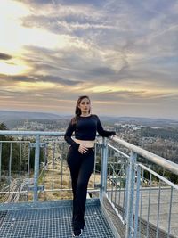 Rear view of woman standing on railing against sky during sunset