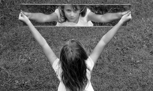 Girl with arms raised holding mirror with reflection on grass