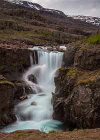 Scenic view of long-exposure waterfall cutting though rocky canyon with snowy mountains behind