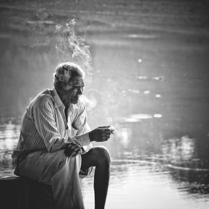 Senior man smoking cigarette while sitting against lake