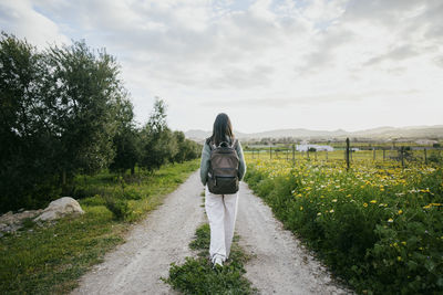 Full length rear view of woman with backpack walking on road by field against sky