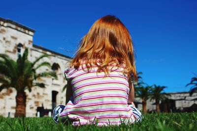 Rear view of girl sitting on grass against blue sky