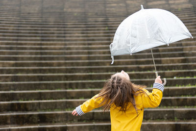 Back view of playful kid in yellow raincoat and with wet umbrella standing on street with outstretched arms and enjoying rainy weather in city