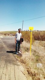Young man reading sign board on grassy field by road against clear blue sky