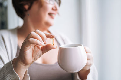 Close up photo of middle aged woman holding omega 3 capsule and mug of water in hands near window 