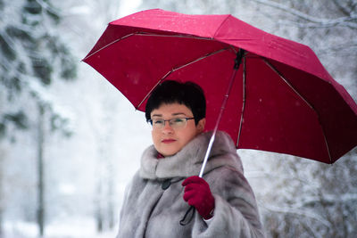 Woman in a gray fur coat with a red umbrella stands on a white, snow-covered background.