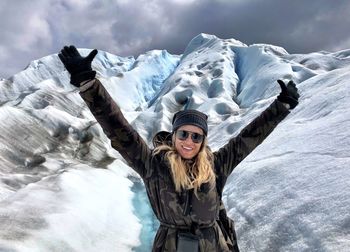 Portrait of smiling woman with arms raised standing on snowcapped mountains during winter