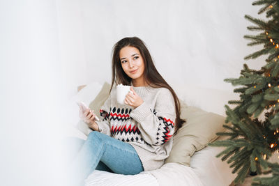 Young asian woman in cozy sweater using mobile smartphone on bed in room with christmas tree at home