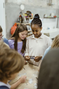 Female students holding electrical part while standing with friends in workshop