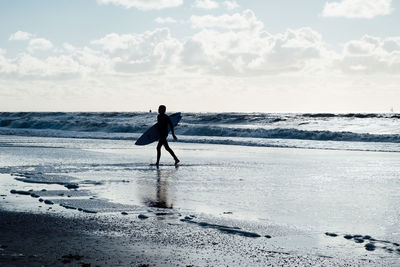 Full length of silhouette surfer walking on shore at beach against sky