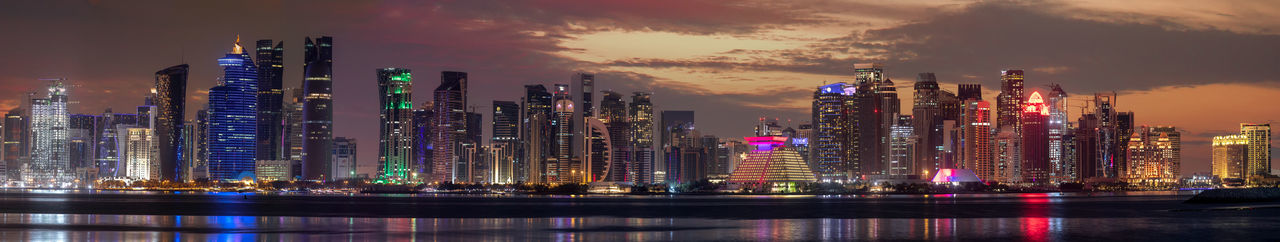 Illuminated modern buildings by river against sky at night