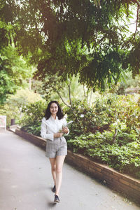 Optimistic asian girl in white shirt and shorts in a park, smiling, walking, carrying laptop and