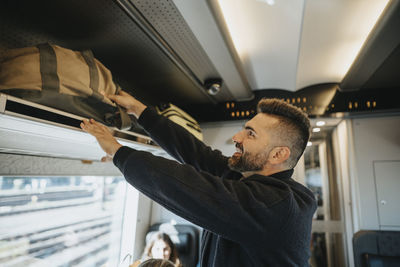 Side view of smiling man loading luggage on shelf in train