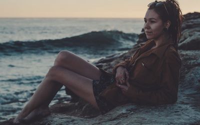 Young woman sitting on shore at beach against sky during sunset