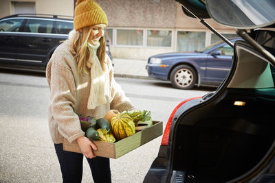 Happy woman with freshly produce vegetables in container standing near car trunk