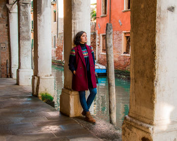 Full length of thoughtful woman looking away while standing against architectural column by canal