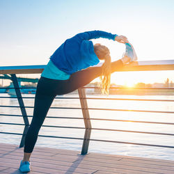 Young woman exercising on promenade by river against clear sky during sunset