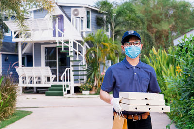 A male delivery man holds a box of goods and food for a customer in front of the house.