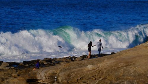 Couple walking on rock while sea waves rushing towards shore