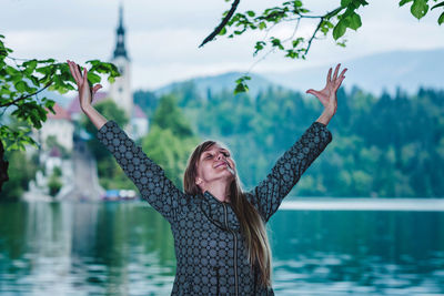 Young woman with arms raised standing by lake against trees