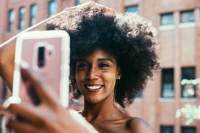 Happy young woman with afro hairstyle taking selfie through smart phone against building