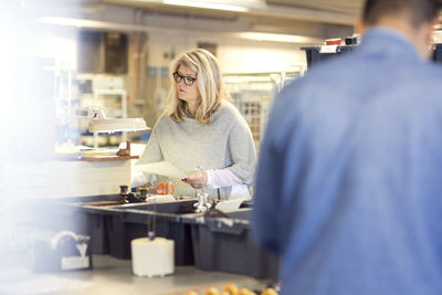 Woman reading paper while volunteer standing by production line at workshop