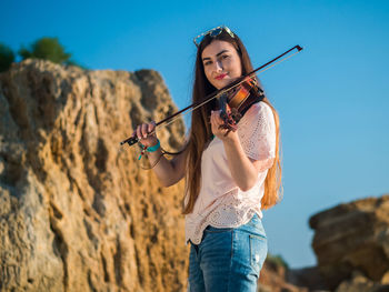 Portrait of young woman playing violin while standing against blue sky