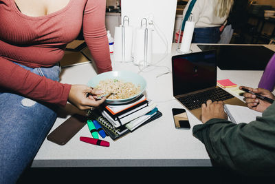 Midsection woman having noodles while male friend using laptop in college dorm