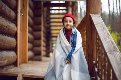 Boy child in a blue denim jacket and hat is sitting at a wooden house