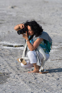 Woman photographing with camera while crouching at beach