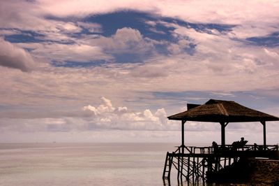 Thatched hut by sea against cloudy sky