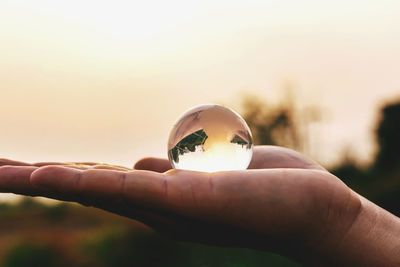 Close-up of hand holding crystal ball against sky during sunset