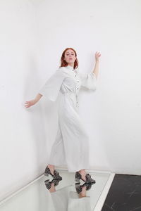 Full length fashion catalog style portrait of woman in jumpsuit standing on mirror over white wall