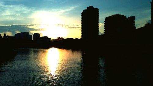 Silhouette buildings by river against sky during sunset