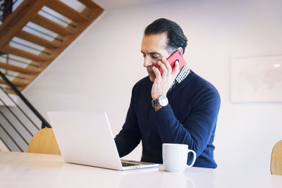 Serious man talking on smart phone while looking at laptop computer at table