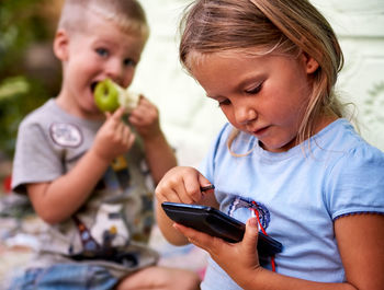 Close-up of girl using phone while sitting with brother outside