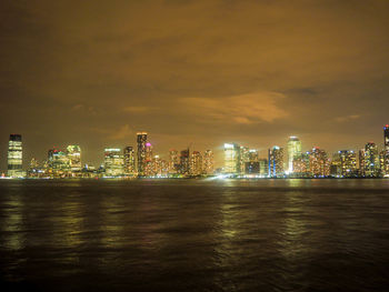 Illuminated city at waterfront against cloudy sky