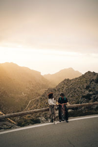 Male and female friends sitting on railing near mountains at sunset