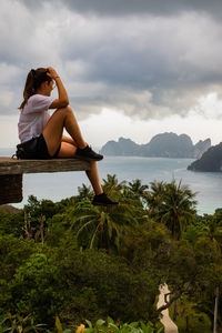 Side view of woman sitting at observation point with sea in background against sky