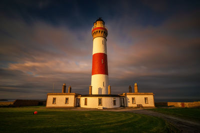 Lighthouse on field by building against sky during sunset