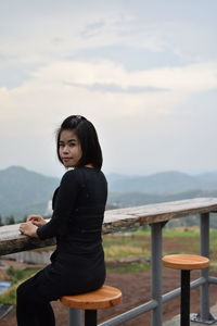 Portrait of young woman sitting on observation point against cloudy sky