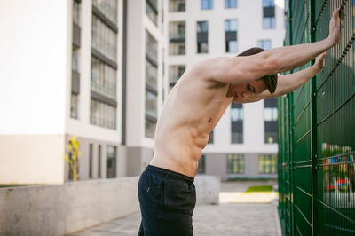 Side view of shirtless man by fence against building