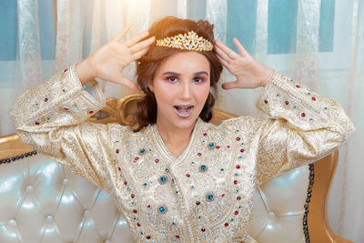 Portrait of young woman wearing crown sitting against wall