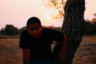 Portrait of man standing by tree trunk against sky during sunset