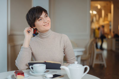 Smiling woman holding book looking away while sitting at cafe