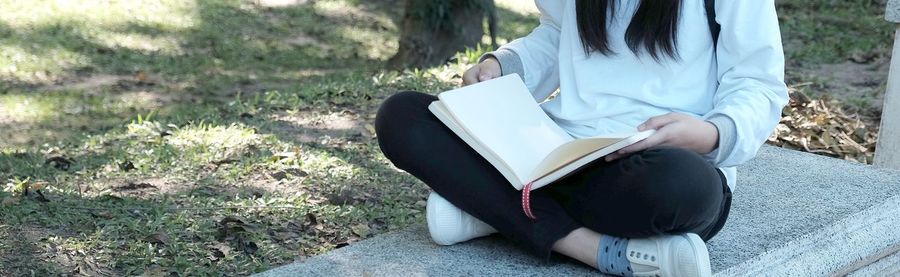 Low section of woman sitting with book on retaining wall at park