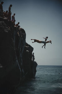 People jumping on rock formation in sea against sky
