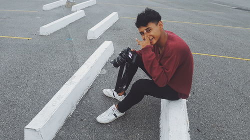 Portrait of smiling man with camera gesturing while sitting on barrier