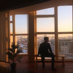 Man sitting on bench against windows during sunset